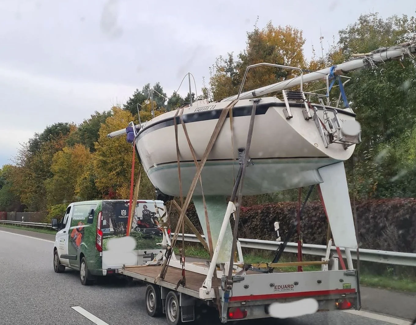 Sailboat secured on Eduard trailer being transported on highway with service vehicle.